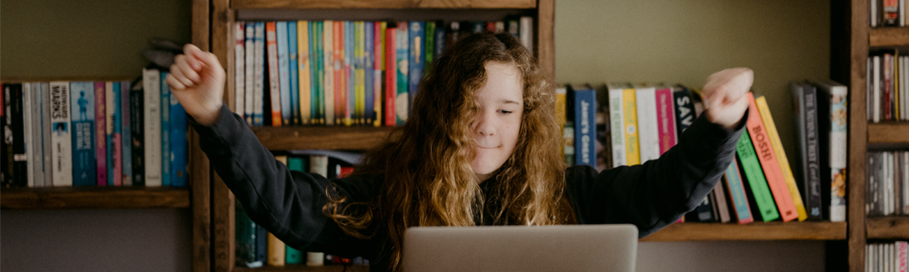 young teen celebrates with hands raised while using laptop in library